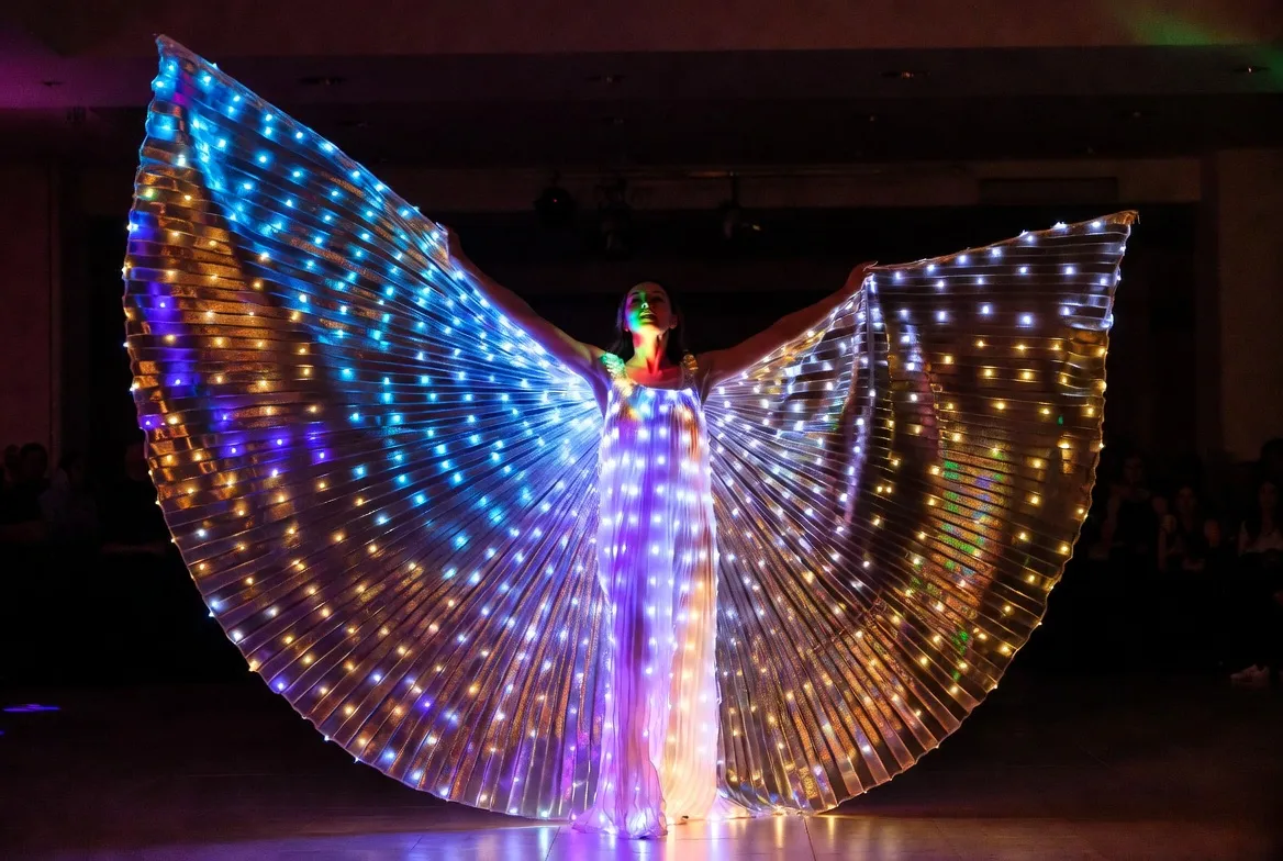 רקדנית עם כנפי LED זוהרות בכחול-סגול-זהב על רחבת ריקודים חשוכה